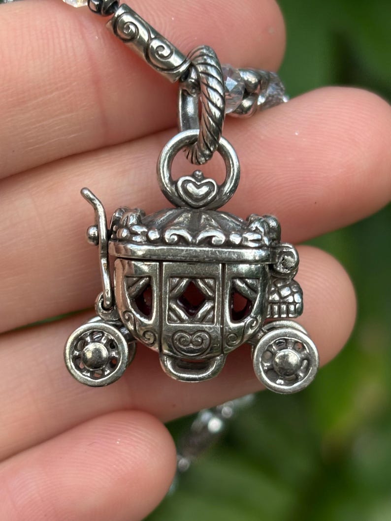 May include: A silver-colored carriage pendant with intricate detailing, including heart-shaped accents and decorative wheels. The carriage windows reveal a red interior. The pendant is suspended from a beaded chain, creating a whimsical accessory.
