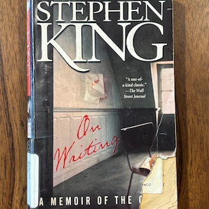 May include: A paperback book titled On Writing: A Memoir of the Craft by Stephen King. The cover features a dark room with a chair and the text The Acclaimed New York Times Bestseller and A one-of-a-kind classic. The bottom right corner is torn.