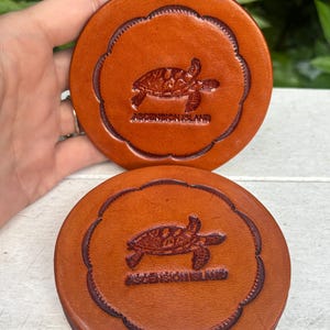 May include: Two round, brown leather coasters. Each coaster features a detailed embossed sea turtle design within a scalloped border. The text "ASCENSION ISLAND" is below the turtle. The coasters are a warm, earthy tone.