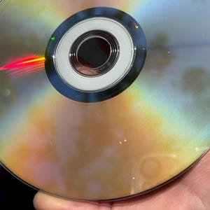 May include: A silver DVD disc with rainbow-like reflections on the surface. The disc has a central hole and a silver ring around it.