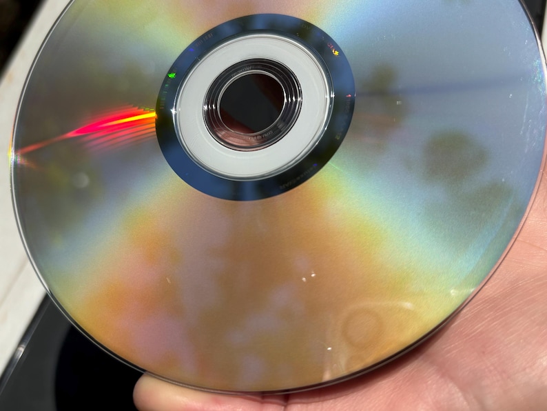 May include: A silver DVD disc with rainbow-like reflections on the surface. The disc has a central hole and a silver ring around it.