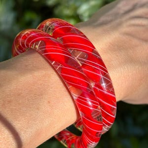 May include: Two red and white striped glass bangles. The bangles are stacked on top of each other and are worn on a person's wrist.