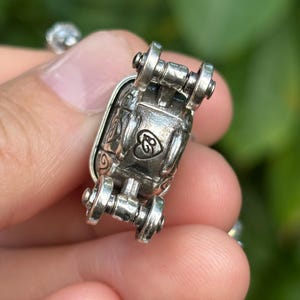 May include: Close-up of a silver-toned jewelry clasp with decorative scrollwork and a heart-shaped design. The clasp has a hinged design with small wheel-like details on each end. The background is blurred, suggesting an outdoor setting.