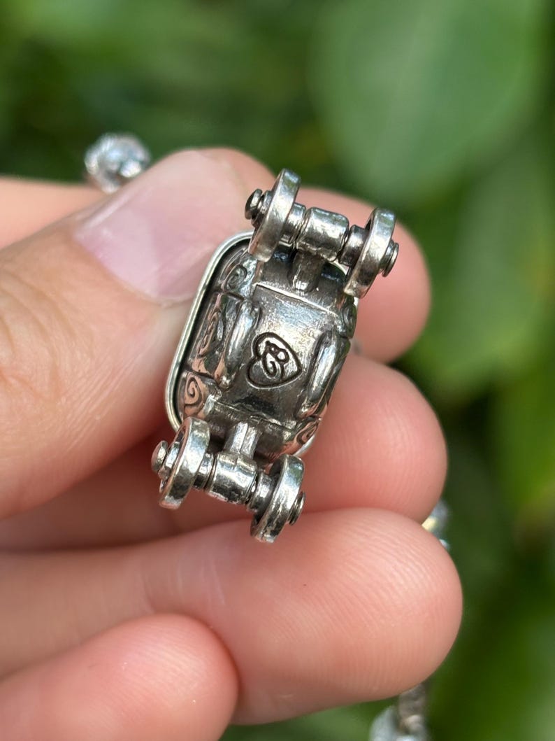 May include: Close-up of a silver-toned jewelry clasp with decorative scrollwork and a heart-shaped design. The clasp has a hinged design with small wheel-like details on each end. The background is blurred, suggesting an outdoor setting.