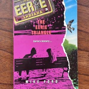 May include: A paperback book titled "The Eerie Triangle" by Mike Ford. The cover features a pink and black design with the words "EERIE INDIANA" and a keyhole graphic. A torn edge reveals a blue sky and a bird.