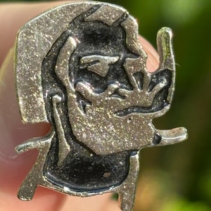 May include: A silver-toned metal pin featuring a stylized profile of a figure with a prominent nose and a textured surface. The pin has a dark, contrasting background within the design, and is held by a person's fingers.