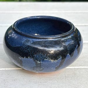 May include: A small, round ceramic bowl with a glossy finish. The bowl features a gradient of dark blue to black hues, with lighter blue accents near the base. The interior is a deep, solid blue. This decorative bowl is suitable for various uses.