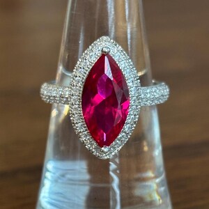 May include: A silver ring with a large, marquise-cut, bright pink gemstone. The gemstone is surrounded by a halo of smaller, clear stones. The ring's band is also encrusted with smaller, clear stones, creating a sparkling effect.