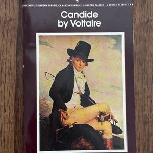 May include: A paperback book titled "Candide by Voltaire" with a portrait of a man in a black coat and hat. The book has a dark burgundy cover with white text and a rooster logo. The text "A Bantam Classic" is printed at the top.