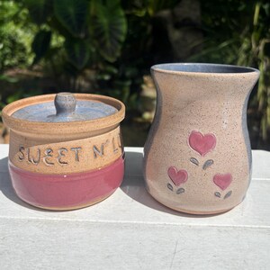 May include: Two handmade ceramic pieces. A lidded jar with the text "SWEET N' LOW" and a vase with three heart designs. The jar is tan, red, and blue. The vase is tan, pink, and blue. Both are on a white surface.