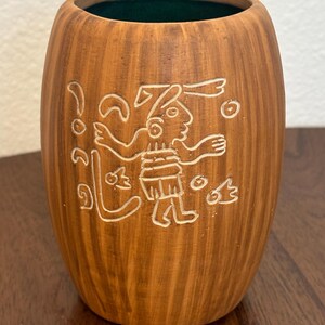 May include: A brown ceramic vase with a carved design of a figure with outstretched arms and other symbols. The vase has a textured surface and a dark green interior. The design is in white.