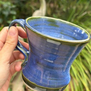 May include: A handmade ceramic mug with a blue glaze and a green rim. The mug has a curved handle and a unique shape. The mug is held in a hand, with a blurred green background.