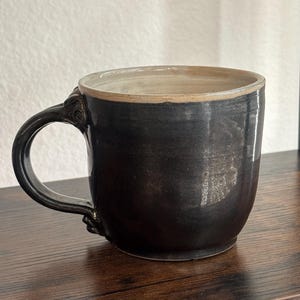 May include: A handmade black ceramic mug with a decorative handle. The mug has a wide, rounded body and a light beige rim. The handle features a detailed, textured design. This mug is perfect for coffee or tea.