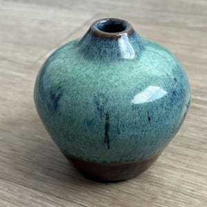 May include: A small, round ceramic vase with a narrow neck. The vase has a glossy finish with a blend of teal and blue hues, and a dark brown base. The interior of the neck is dark brown.
