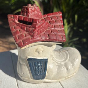 May include: A white cast iron piggy bank shaped like a boot with a red brick house on top. The house has a chimney and a slot for coins. The boot has a blue door with a white frame.
