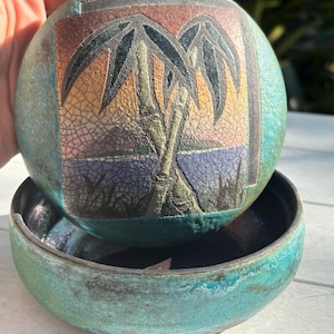 May include: A teal ceramic trinket box with a lid. The lid features a square design with a hand-painted image of palm trees and a sunset. The box is perfect for storing small items like jewelry or trinkets.
