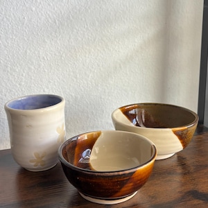 May include: A ceramic cup with a light exterior, pale flower patterns, and a lavender interior, accompanied by two bowls featuring a two-tone design with a glossy dark brown glaze on one side and a smooth cream-colored finish on the other side.