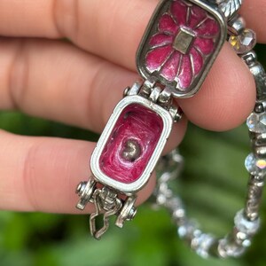 May include: A silver-toned locket bracelet with a hinged rectangular charm. The charm opens to reveal a pink interior and a small object. The exterior of the charm features a pink floral design. The bracelet has a beaded chain.