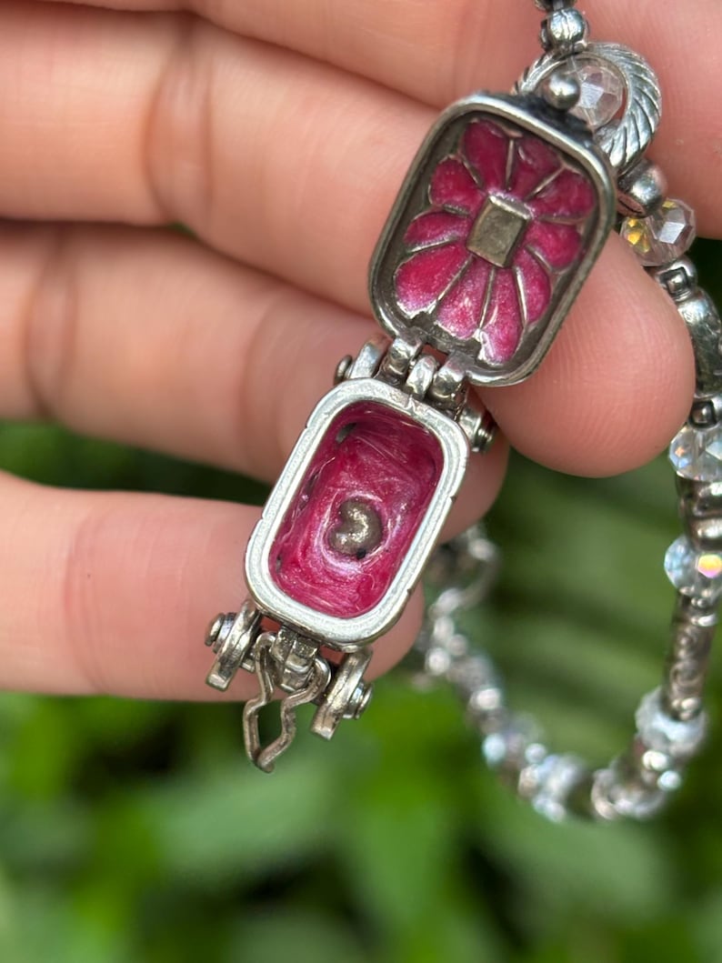 May include: A silver-toned locket bracelet with a hinged rectangular charm. The charm opens to reveal a pink interior and a small object. The exterior of the charm features a pink floral design. The bracelet has a beaded chain.