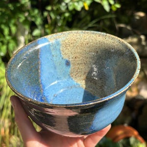 May include: Handmade ceramic bowl with a glossy finish. The bowl features a gradient of blue and tan hues, with a darker blue rim. The interior has a speckled, textured appearance. A unique piece for serving or display.