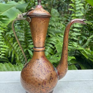May include: A vintage copper pitcher with a long, curved spout and a hinged lid. The pitcher has a hammered texture and a handle. The copper has a warm, reddish-brown hue, with some areas showing darker patina.