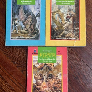 May include: Three paperback books with red, yellow, and blue covers. Each book features a dragon illustration and the title "Dragonfire". The titles of the books are "Thieves of Nir", "Castle Beneath The Sea", and "The Curse of Peredur".