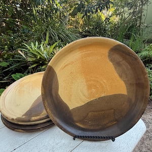 May include: Set of four ceramic plates with a brown and yellow glaze. The plates are stacked on a white wooden table.
