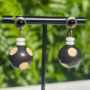 May include: A pair of dangle earrings. Each earring features a black stud with a small black stone, connected to a white bead stack, and a large black sphere with cream-colored polka dots. The earrings are displayed on a black stand.
