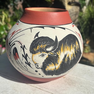 May include: Hand-painted ceramic pot with a cream-colored body and a red rim and base. The pot features a detailed illustration of a bison in black, yellow, and white, with decorative tribal-inspired elements.
