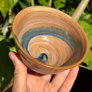 May include: Handmade ceramic bowl with a spiral design. The bowl features a brown exterior with a blue and brown interior. The rim is a darker brown. The bowl is held in a hand against a background of green leaves.