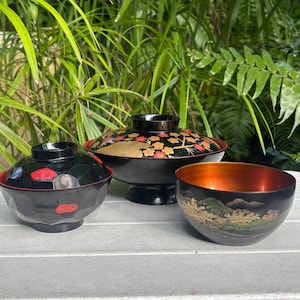 May include: Three Japanese lacquerware bowls. The bowls are black with red and gold accents. One bowl has a lid with a floral design. The other bowl has a landscape design. The bowls are on a white surface.
