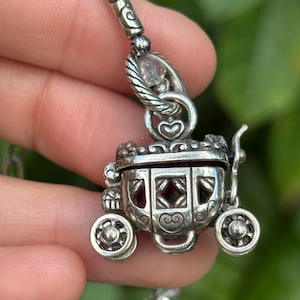 May include: A silver-toned carriage pendant with intricate detailing, including heart-shaped accents and decorative wheels. The pendant is suspended from a beaded chain with clear crystal beads. The carriage has a hinged top and a red interior.