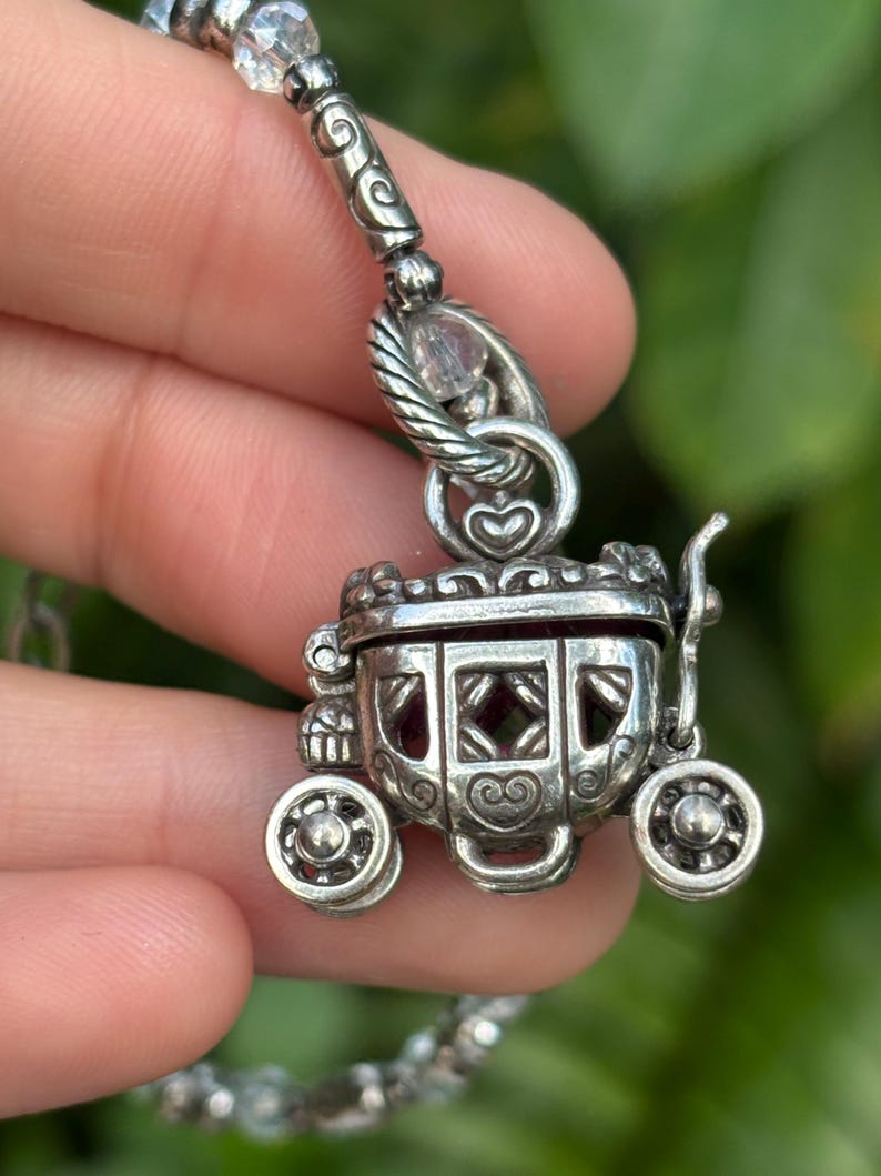 May include: A silver-toned carriage pendant with intricate detailing, including heart-shaped accents and decorative wheels. The pendant is suspended from a beaded chain with clear crystal beads. The carriage has a hinged top and a red interior.