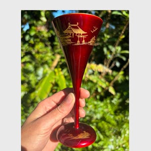 May include: A red glass goblet with a gold design of a Japanese temple and trees. The goblet is held in a hand against a green leafy background.