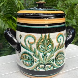 May include: A ceramic pot with a lid, featuring a black, yellow, and teal striped design on the lid and rim. The body is white with green and yellow floral patterns and black handles. A decorative kitchenware item.