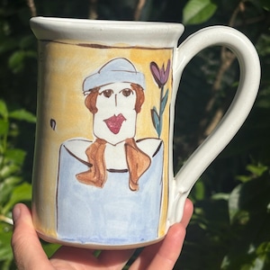 May include: A white ceramic mug with a hand-painted illustration of a woman in a blue hat and dress, with brown hair and a red mouth. The mug has a yellow background and a purple flower. The handle is white.