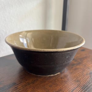 May include: A round ceramic bowl featuring a dark charcoal exterior and a smooth, light beige interior glaze. The rim of the bowl is unglazed, revealing a natural tan texture that contrasts with the dark base and the glossy interior surface.