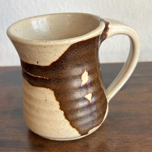 May include: A ceramic mug with a speckled cream-colored base and a wide, dark brown vertical stripe. The mug features a flared rim, a curved handle, and a series of horizontal ridges along the lower body, creating a textured surface finish.