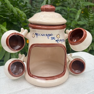 May include: Cream-colored ceramic container with a brown rim and lid, featuring five attached cup-like structures. The text "Recuerdo de Merida" is written on the front. The container is likely for decorative or functional use.