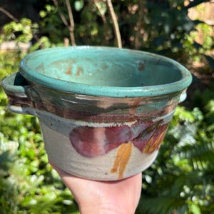 May include: A handmade ceramic planter with a teal glaze and a brown, white, and purple design. The planter has two handles on the sides.
