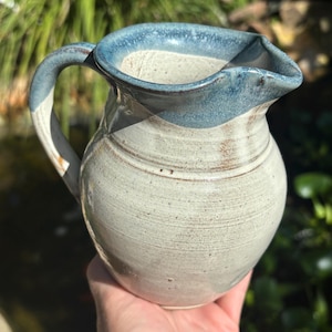 May include: Handmade ceramic pitcher with a rounded body and a curved handle. The pitcher features a light gray base with a blue rim and handle accents. The pitcher is held in a hand, with a blurred green background.