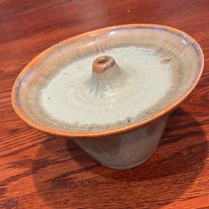 May include: A ceramic incense burner with a light green glaze and a brown rim. The burner has a small, cone-shaped opening in the center.