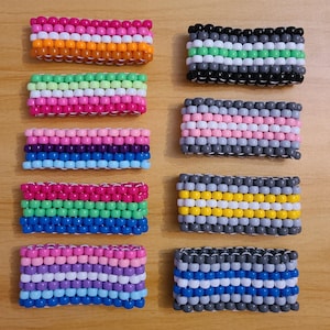 May include: Ten colorful beaded bracelets with different pride flag designs. The bracelets are made with small, round beads in various colors, including pink, blue, green, yellow, white, and black. The bracelets are arranged in two rows of five, with each row representing a different flag.