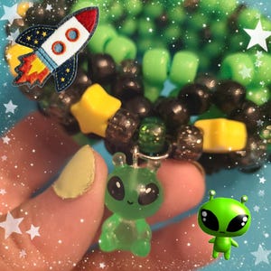 Op de afbeelding: Een handgemaakte kandi-armband met groene, zwarte en gele kralen. De armband heeft een groene alien-bedel en een raketpatch. De armband is een leuk accessoire voor raves of festivals.