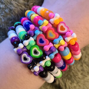 May include: A collection of colorful beaded bracelets. The bracelets feature a variety of shapes, including hearts and bone-shaped beads, in shades of pink, blue, green, yellow, and purple. The bracelets are stacked on top of each other.