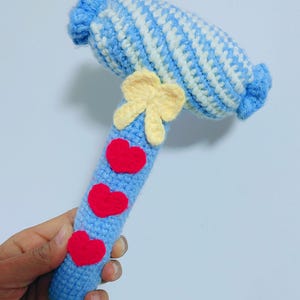 May include: A handmade crochet hammer toy in shades of blue and white, with a yellow bow and three red hearts. The hammer head has a striped pattern, and the handle is adorned with hearts.