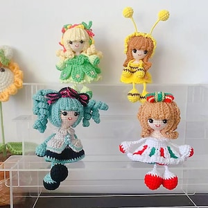 Könnte beinhalten: Vier gehäkelte Puppen mit verschiedenen Outfits und Frisuren. Die Puppen sitzen auf einem durchsichtigen Acrylregal. Die Puppen tragen ein grünes Kleid, ein gelbes und schwarzes Bienenkostüm, ein blaues und weißes Kleid und ein rotes und weißes Kleid.
