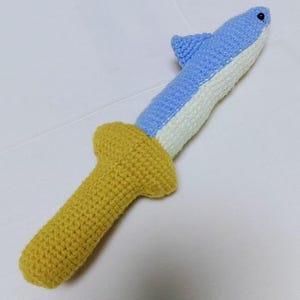 Könnte beinhalten: Ein handgefertigtes Häkeltier in Form eines Haifisch-Schwerts. Der Hai ist blau und weiß, mit einem gelben Griff. Das Spielzeug ist aus weichem Garn gefertigt und hat ein skurriles Design.