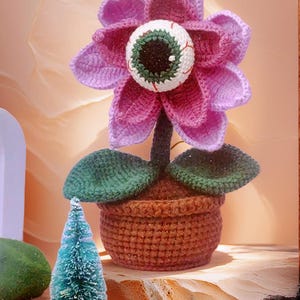 May include: A crocheted flower with a large eye in the center, in a brown pot with green leaves. The flower is pink and purple, and the eye is green and white.