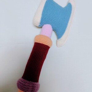 May include: A handmade crochet axe toy. The axe head is light blue with a white border. The handle is burgundy with a pink and orange section, and a purple and orange base. This soft toy is suitable for children.